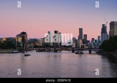 Panorama crépusculaire des gratte-ciel de Londres sur la Tamise avec pont illuminé et bateaux reflétant le ciel rose violet couché de soleil sur les gratte-ciel modernes Banque D'Images