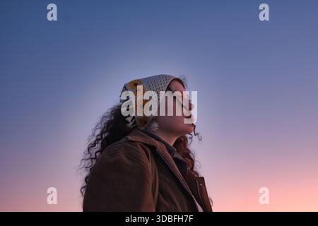 Jeune femme de profil portant un foulard crochet tournesol avec boucles d'oreilles Daisy contre gradient Blue Sky au coucher du soleil Banque D'Images