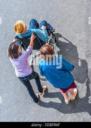 Vue aérienne de deux femmes aides poussant un homme plus âgé invalide dans un fauteuil roulant - centre de la France. Banque D'Images