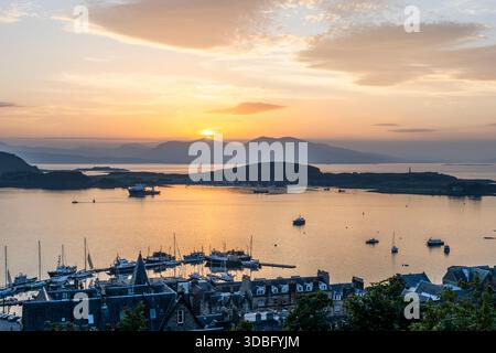 Coucher de soleil doré sur la baie d'Oban, silhouettes de bateaux à proximité et une ville côtière pittoresque au premier plan. L'eau calme reflète des teintes chaudes comme dis Banque D'Images