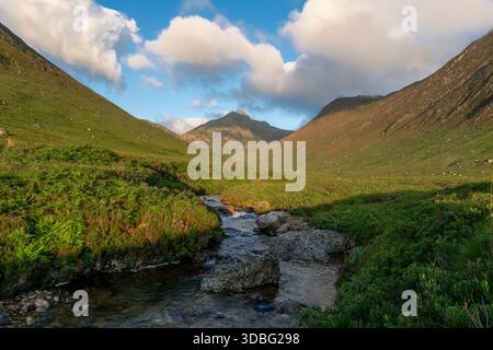 Un paysage serein avec des collines verdoyantes, un ruisseau sinueux et des montagnes escarpées sous un ciel bleu vif avec des nuages moelleux ; idéal pour la nature, Banque D'Images