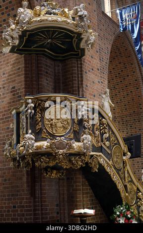 Vue latérale d'une chaire baroque richement décorée avec des sculptures dorées, des panneaux en relief et des figures sculptées sur les murs de briques rouges de l'église. Banque D'Images