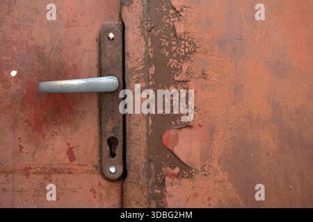 Gros plan du panneau de porte en métal altéré avec poignée grise, plaque de verrouillage rouillée, couches de peinture marron et rouge écaillées, textures de corrosion visibles. Banque D'Images