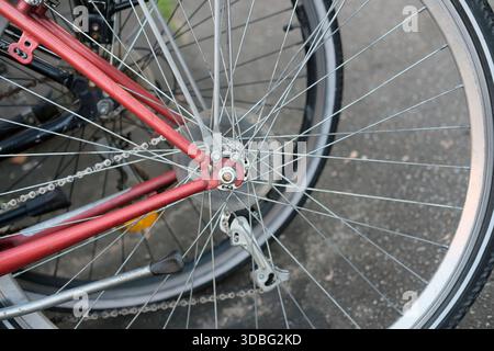 Gros plan de la roue arrière d'un vélo de ville rouge avec rayons métalliques, chaîne, frein de moyeu et béquille, photographié sous angle latéral en lumière naturelle. Banque D'Images