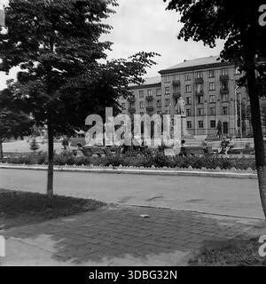 Photo d'archives en noir et blanc capturant une scène paisible de parc urbain dans la région de Donetsk (probablement Druzhkivka), Ukraine, Donbass, années 1960 Une statue de Vladimir Lénine se dresse sur un piédestal entouré de parterres de fleurs et de bancs occupés par des citoyens au repos. En arrière-plan, un grand bâtiment résidentiel en briques (architecture 'stalinienne') est visible. Les femmes à la mode vintage se promènent le long de la passerelle pavée. Une vision nostalgique de la vie quotidienne urbaine soviétique et des loisirs dans l'ère d'avant-guerre. Banque D'Images