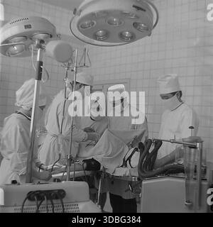 Photo d'archives en noir et blanc capturant une équipe de chirurgiens effectuant une opération complexe dans un hôpital de Donetsk, Ukraine, Donbass, années 1960 Le personnel médical en chapeaux blancs et masques travaille avec une concentration intense sous une grande lampe sans ombre. Au premier plan, une machine d'anesthésie vintage avec des tubes en verre surveille le patient. Un aperçu puissant du système de santé avancé, du devoir professionnel et du sauvetage de vies dans le centre scientifique de la région pacifique bien avant la guerre. Banque D'Images