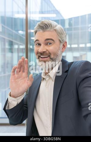 Homme d'affaires senior avec une barbe grise et une tenue professionnelle souriant joyeusement tout en agitant une main amicale à la caméra, debout dans un environnement de bureau moderne Banque D'Images
