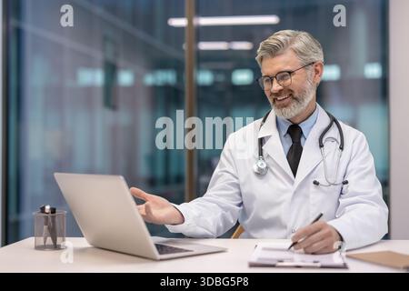 Un médecin masculin souriant portant une blouse de laboratoire blanche et un stéthoscope a un appel vidéo sur un ordinateur portable tout en faisant des gestes et en prenant des notes, fournissant une assistance médicale à distance Banque D'Images
