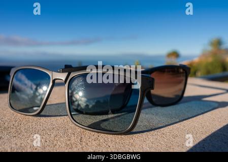 Deux paires de lunettes de soleil noires encadrées reposent sur une pierre ensoleillée à Funchal, Madère. L'horizon océanique et les palmiers s'estompent. Les reflets montrent des nuages en lumière vive Banque D'Images