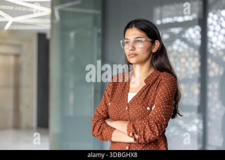 Jeune femme d'affaires indienne adulte portant des lunettes et une chemise à pois, debout avec les bras croisés, regardant soigneusement loin dans un bureau moderne, représentant la confiance et le succès Banque D'Images