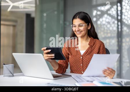 Jeune femme travaillant sur les finances et la comptabilité, tenant une calculatrice et des documents, tout en souriant et en regardant un ordinateur portable dans un environnement de bureau professionnel lumineux Banque D'Images