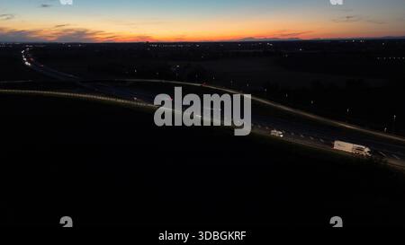 Vue aérienne de nuit montrant le trafic dense sur l'autoroute A21 Piacenza Brescia pendant le coucher du soleil, voitures et camions se déplaçant le long éclairant la route avec th Banque D'Images