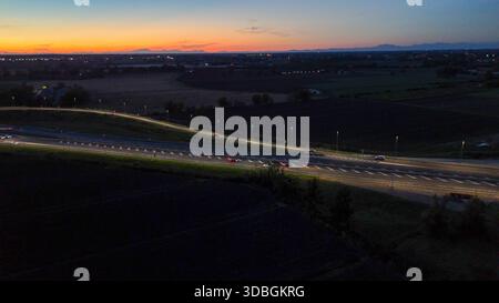 Vue aérienne de nuit montrant le trafic dense sur l'autoroute A21 Piacenza Brescia pendant le coucher du soleil, voitures et camions se déplaçant le long éclairant la route avec th Banque D'Images