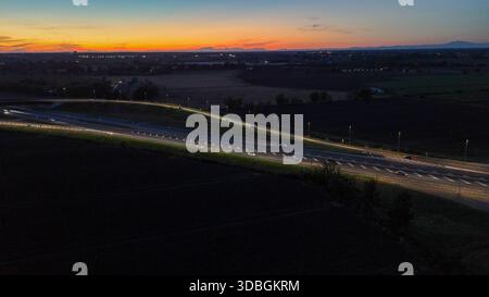 Vue aérienne de nuit montrant le trafic dense sur l'autoroute A21 Piacenza Brescia pendant le coucher du soleil, voitures et camions se déplaçant le long éclairant la route avec th Banque D'Images