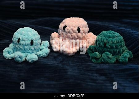 Trois peluches de poulpe crochetées à la main en bleu pastel, rose doux et vert foncé reposent sur un fond de tissu texturé foncé. Jouets mignons amigurumi. Banque D'Images