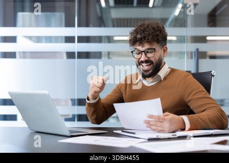 Jeune homme recevant de bonnes nouvelles au travail, démontrant la réussite et le succès avec une expression joyeuse et un geste triomphant tout en travaillant sur un ordinateur portable à un bureau contemporain Banque D'Images