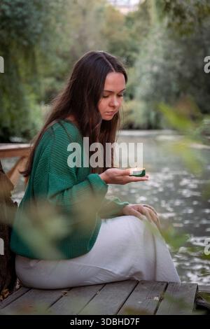 Jeune femme dans un pull vert tient une petite bougie allumée tout en étant assise près d'un étang calme, lumière du jour douce, tons verts muets, humeur calme et contemplative Banque D'Images