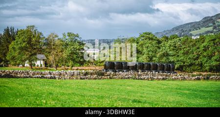 Vue panoramique téléphoto de la campagne rurale avec prairie verte, mur de pierre et balles empilées d'ensilage noir, arbres et collines sous ciel nuageux dramatique, Banque D'Images