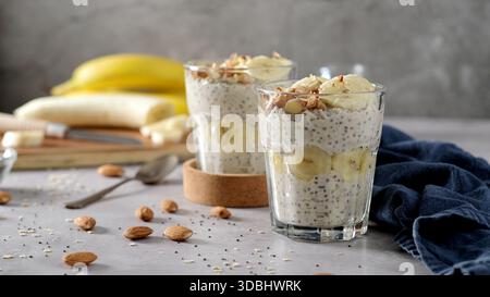 Deux tasses en verre remplies de pudding crémeux aux graines de chia recouvertes de tranches de bananes fraîches et recouvertes d'amandes écrasées. La scène lumineuse et minimaliste fea Banque D'Images