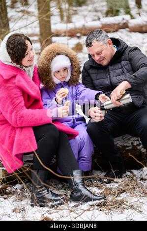 Aventures hivernales en famille dans la nature : les parents et la fille apprécient une collation confortable à l'extérieur. Maman, papa, fille sirotent une boisson chaude du thermos tout en étant assis sur le rondin pendant la promenade en forêt, en dégustant une collation. Promenade hivernale Banque D'Images
