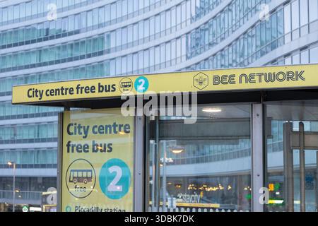 Manchester, royaume-uni, 19 janvier 2025 panneau d'arrêt de bus jaune vif avec un badge bleu '2', annonçant un service de bus gratuit. Installé contre un bâtiment en verre élégant, Banque D'Images