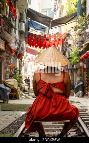 Vue d'une femme dans une robe rouge vif et chapeau conique traditionnel se trouve sur les voies ferrées sous des lanternes rouges festives, Hanoi, Hanoi, Vietnam. Banque D'Images