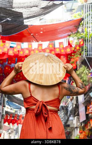 Vue de lanternes rouges vibrantes et de décorations bordent les rues animées comme une femme dans une robe orange pose avec un chapeau conique, Hanoi, Hanoi, Vietnam. Banque D'Images
