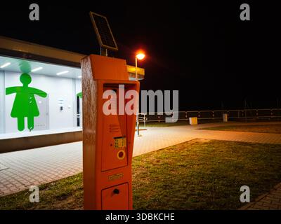 Photo nocturne d'un pilier de téléphone orange SOS près d'une salle de bains publique avec un signe figuratif vert en Pologne. Banque D'Images