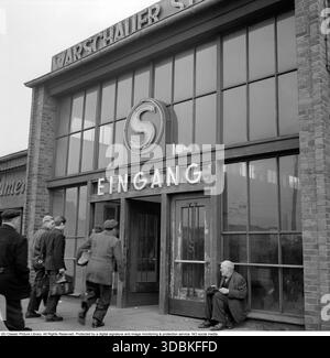 Entrée de la station Warschauer Straße, Berlin est (1957) cette photo de 1957 montre l'entrée extérieure (Eingang) de la station Warschauer Straße S-Bahn (et probablement U-Bahn) à Friedrichshain, Berlin est (Ostberlin). Le panneau lumineux « S » au néon marque le S-Bahn de Berlin – le réseau ferroviaire de banlieue de la ville – tandis que le bâtiment porte le nom de la gare « WarschauerStraße »). Dans la ville divisée de la guerre froide (quatre ans avant le mur de Berlin), il s'agissait d'un centre de transport clé reliant Berlin-est au reste du réseau, toujours exploité par la Reichsbahn est-allemande mais accessible des deux côtés. Les trains traversaient s Banque D'Images