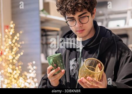 Adolescent choisissant une tasse de Noël et un décor en verre festif pendant les achats de vacances. Jeune homme comparant la tasse des fêtes et l'article en verre décoratif à Christma Banque D'Images