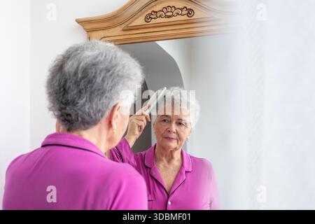 une femme âgée se peigne les cheveux le matin près du miroir. Routine matinale, soins de la peau, anti-âge, soins personnels, beauté senior, confort à la maison, Banque D'Images