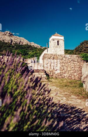 Vue sur un chemin de pierre monte à une chapelle blanche altérée avec un toit en terre cuite sur fond de montagnes escarpées, encadrées par la lavande floue, Kr Banque D'Images