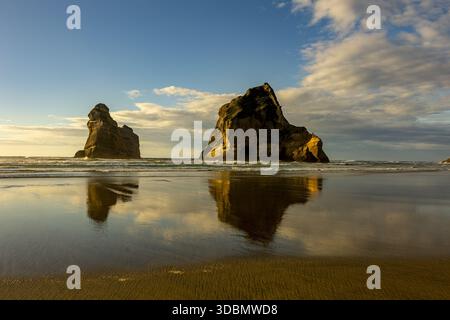 Vue de formations rocheuses monolithiques se dressant contre les vagues qui s'écrasent, reflétées dans le sable humide sous un vaste ciel, plage de Wharariki, Pūponga, T. Banque D'Images