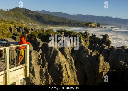 Vue d'une femme en tenue rouge vif regardant depuis une plate-forme en bois au-dessus de rochers déchiquetés et sombres rencontrant l'océan mousseux, Punakaiki, région de Tasman, Nouveau Banque D'Images