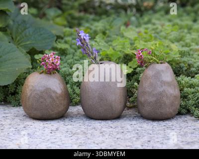 Trois vases en grès faits à la main avec de petites fleurs sauvages disposées à l'extérieur sur la surface de pierre, entourés de feuillage vert. Reflec. Natures mortes à lumière naturelle Banque D'Images