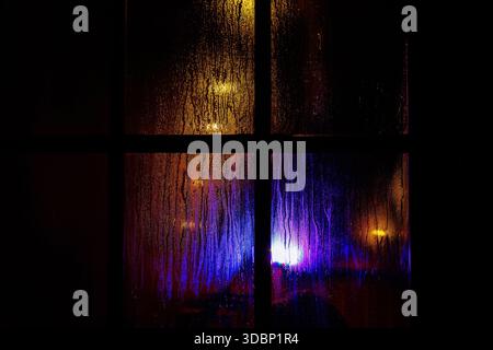 Une vitre sombre avec des gouttelettes de pluie la nuit. Lumières de noël jaunes et bleues floues à travers le verre humide recouvert de gouttes de condensat. Banque D'Images