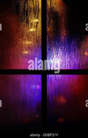 Une vitre sombre avec des gouttelettes de pluie la nuit. Lumières de noël jaunes et bleues floues à travers le verre humide recouvert de gouttes de condensat. Banque D'Images