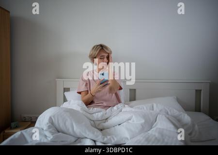 Femme d'âge moyen fatiguée ayant mal à la tête tout en procrastinant avec smartphone au lit le matin. Banque D'Images