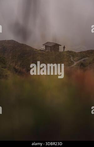 Une cabane en bois solitaire se trouve entre deux collines herbeuses au milieu du paysage alpin des Alpes suisses. Un brouillard mystique obscurcit l'arrière-plan ; au premier plan, un chemin mène à travers la prairie jusqu'à la cabane Banque D'Images