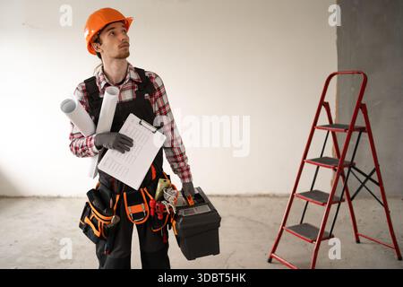 ouvrier masculin ou ingénieur dans le casque et la ceinture à outils tenant la boîte à outils se préparant à travailler dans la nouvelle maison moderne. copier l'espace Banque D'Images