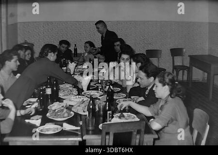 Sloviansk, Ukraine, URSS - années 1950 : photographie d'intérieur d'archive d'un grand banquet festif ou célébration. Un groupe d'hommes et de femmes sont assis à de longues tables en bois remplies de nourriture et de boissons dans une salle simple. Un homme se tient debout pour porter un toast pendant que les invités écoutent et lèvent leurs verres. La scène capture l'esprit communautaire et la vie sociale du peuple soviétique. Cette image historique documente un moment de joie et d’unité dans la région pacifique du Donbass, reflétant la vie quotidienne avant la guerre. Banque D'Images