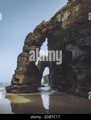 Vue sur les majestueuses formations rocheuses avec leurs arches naturelles reflétées sur le sable humide sous un ciel dégagé, Ribadeo, Lugo, Espagne. Banque D'Images