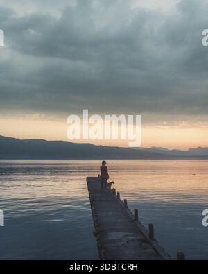 Vue d'une personne debout sur une jetée en bois s'étendant dans le lac tranquille sous un ciel sombre peint avec des teintes orange et grises douces, le lac Windermere, Banque D'Images