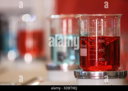 Gros plan précis d'un bécher en verre rempli de liquide rouge à l'échelle du laboratoire, avec d'autres béchers colorés flous en arrière-plan Banque D'Images