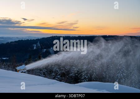 Le canon à neige fonctionne sur une pente de montagne, créant de la neige artificielle pour le ski. Le soleil se couche en arrière-plan, ajoutant de la couleur au paysage hivernal. Les arbres sont couverts de neige. Banque D'Images