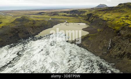 Vue aérienne de glace glaciaire rencontrant un lac chargé de limon, contrasté par des falaises sombres et des plaines lointaines sous un ciel couvert, Solheimajokull, Myrdalshrepp Banque D'Images