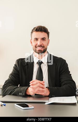 Un homme d'affaires confiant dans un costume formel sourit tout en étant assis à son bureau, faisant preuve de professionnalisme lors d'une réunion de bureau. Il s'engage directement Banque D'Images