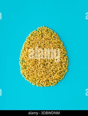 Forme d'oeuf de Pâques faite de pâtes macaroni crues de coude sur fond turquoise. Concept créatif de nourriture de Pâques. Aménagement festif des fêtes. Pâtes esthétiques. Banque D'Images
