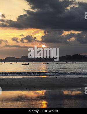 Vue du coucher de soleil sur l'océan, jetant une teinte dorée sur l'eau et le sable sombre, avec des bateaux silhouettés contre l'horizon, Lombok, Ouest Banque D'Images