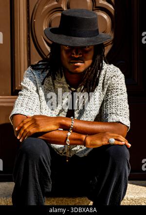 Un homme avec des dreadlocks portant un chapeau noir et une chemise à motifs est assis sur des marches avec les bras croisés. Banque D'Images